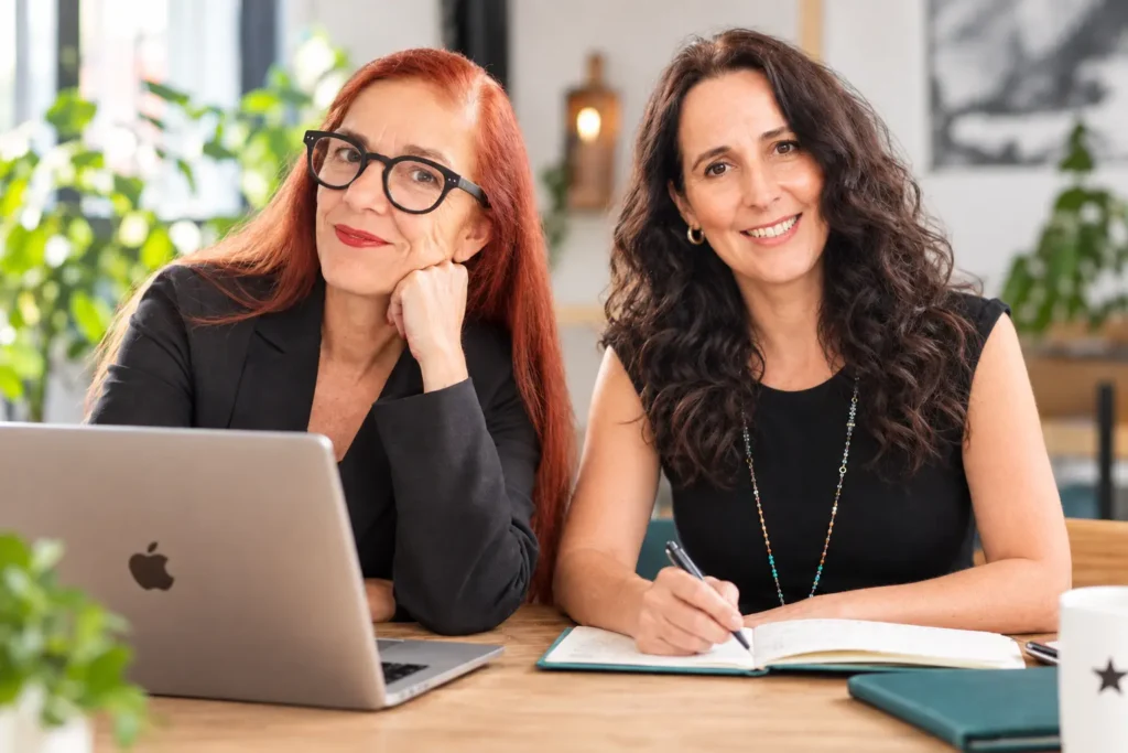 Corinna und Anne von CAJ Marketing sitzen gemeinsam am Schreibtisch mit Laptop und Notizbuch und beraten kleine sowie mittelständische Unternehmen als externe Marketingabteilung.